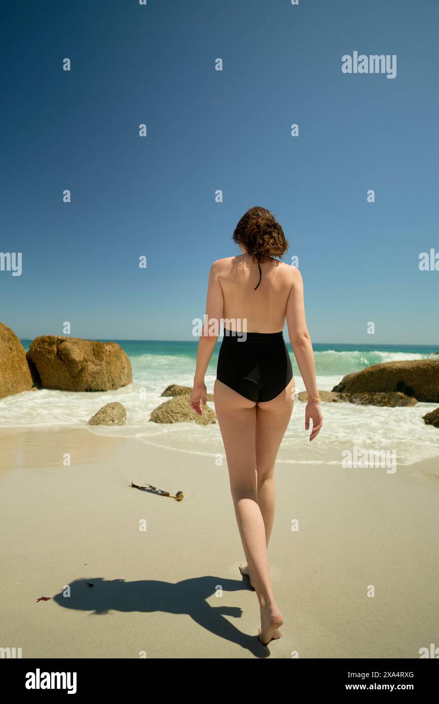 Eine Frau in schwarzem Badeanzug, die an einem Sandstrand unter klarem blauem Himmel zum Meer geht. Stockfoto