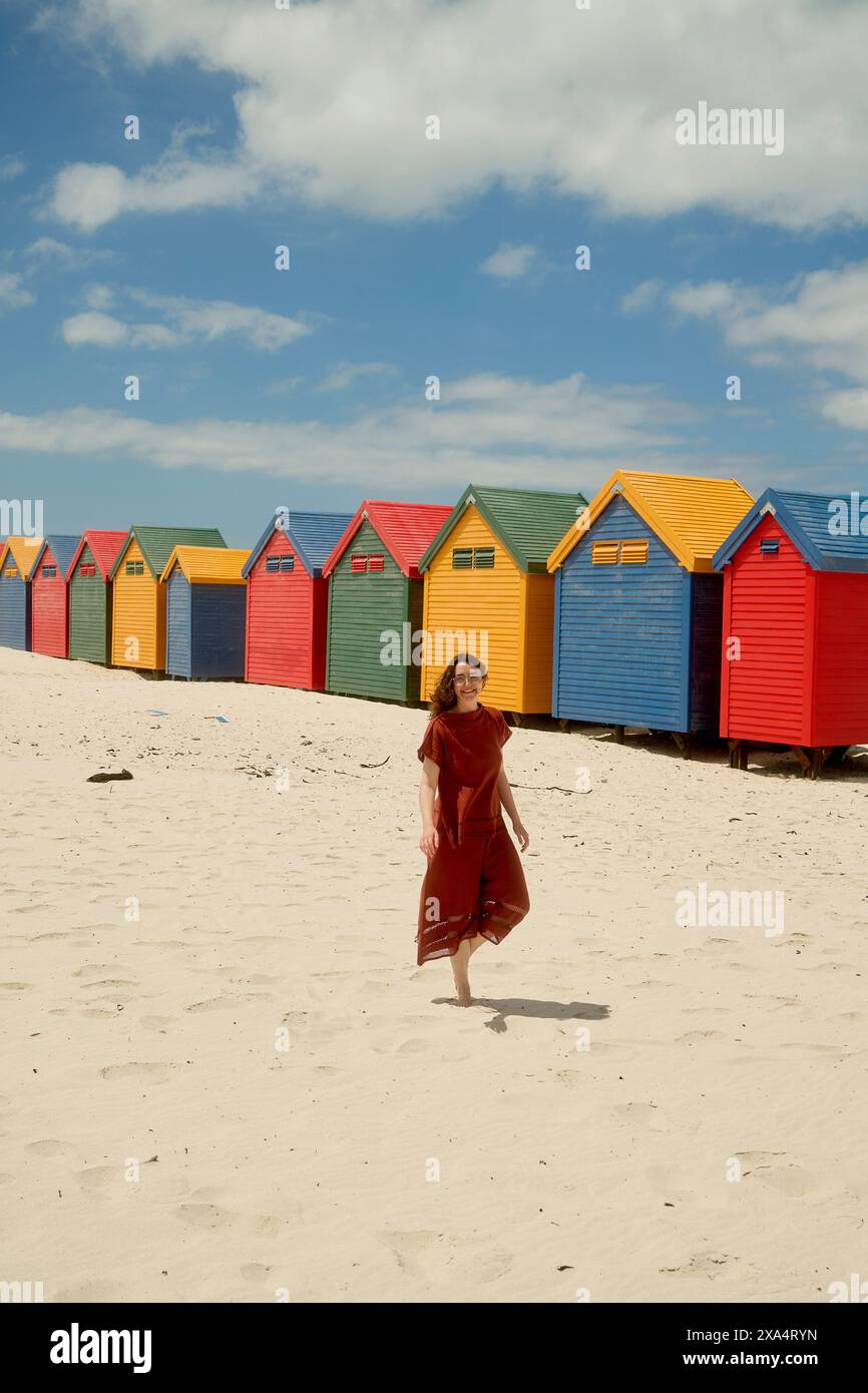 Frau in einem roten Kleid, die einen sonnigen Tag am Strand mit bunten Strandhütten im Hintergrund genießt. Stockfoto