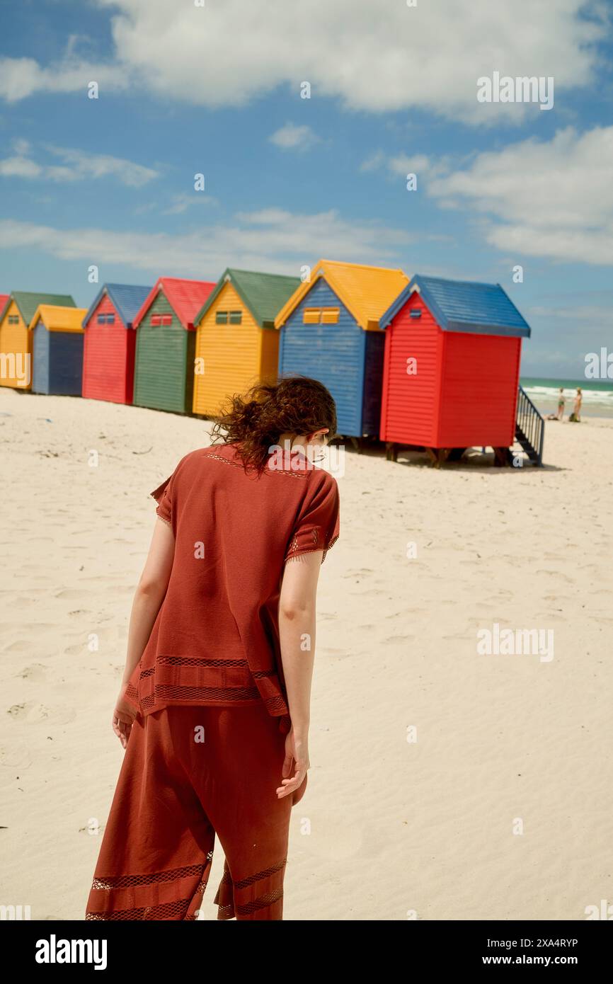 Eine Frau in rotem Outfit steht an einem Sandstrand mit einer Reihe von bunten Strandhütten unter einem bewölkten Himmel im Hintergrund. Stockfoto
