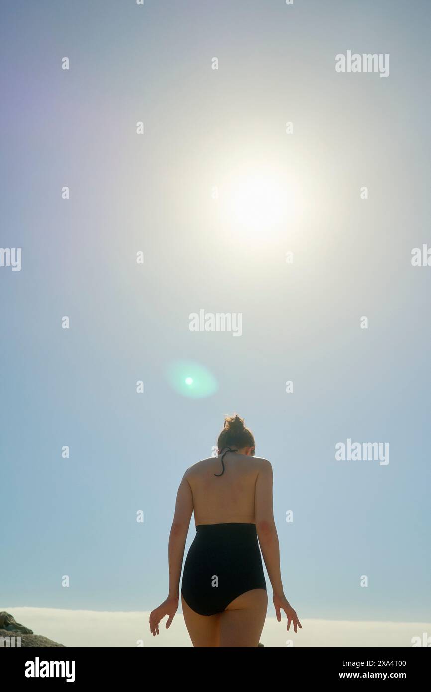 Eine Frau in schwarzem Badeanzug steht auf einem Felsen, der Sonne zugewandt, in einem klaren blauen Himmel. Stockfoto