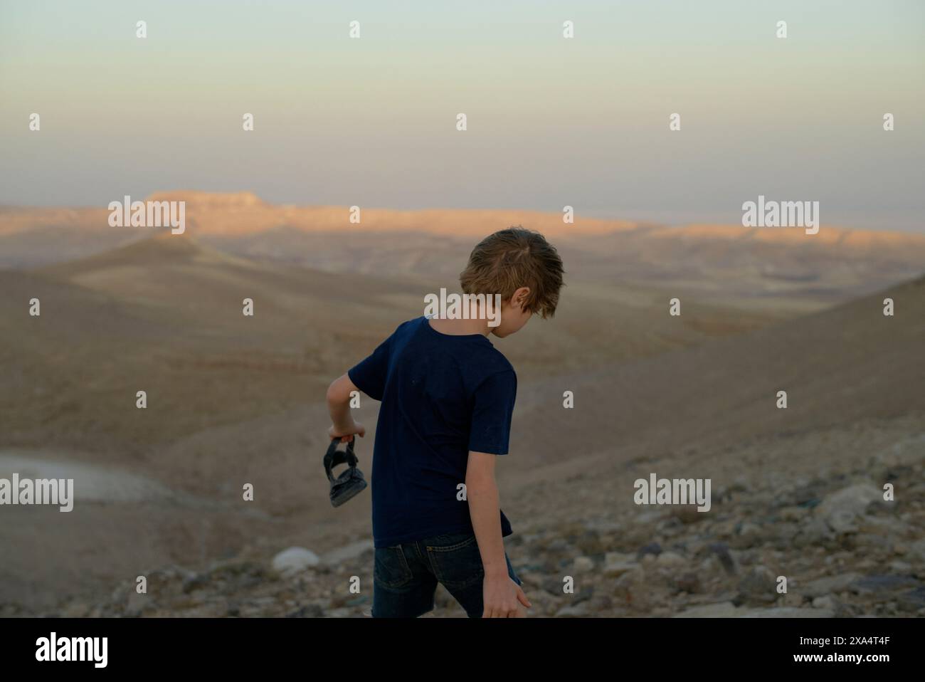 Ein kleiner Junge spaziert in der Abenddämmerung durch eine felsige Wüstenlandschaft mit Bergen im Hintergrund. Stockfoto