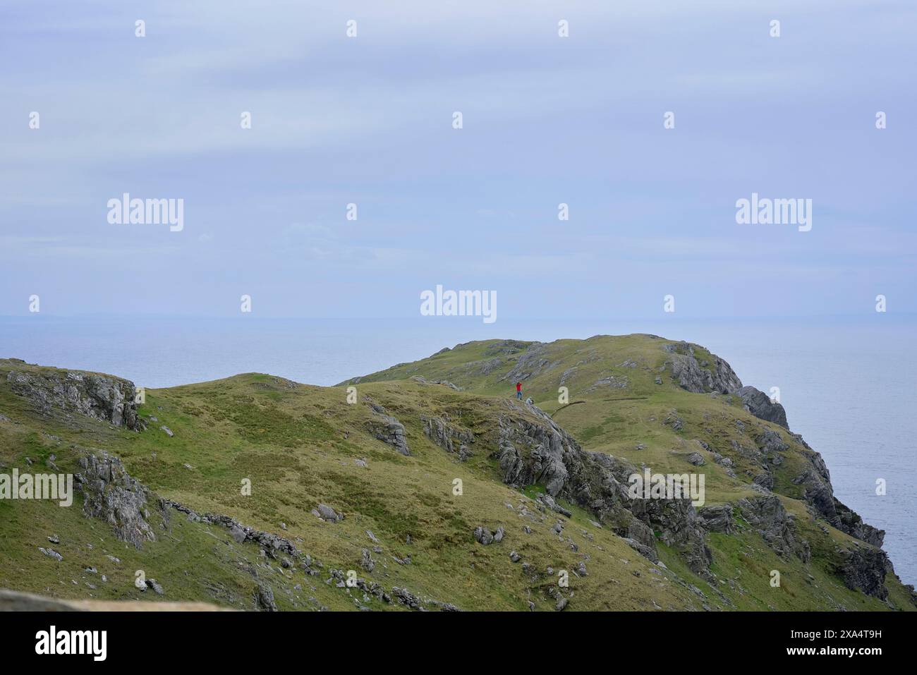 Ein einsamer Wanderer in roter Jacke steht auf einem zerklüfteten Bergweg mit Blick auf das Meer. Stockfoto