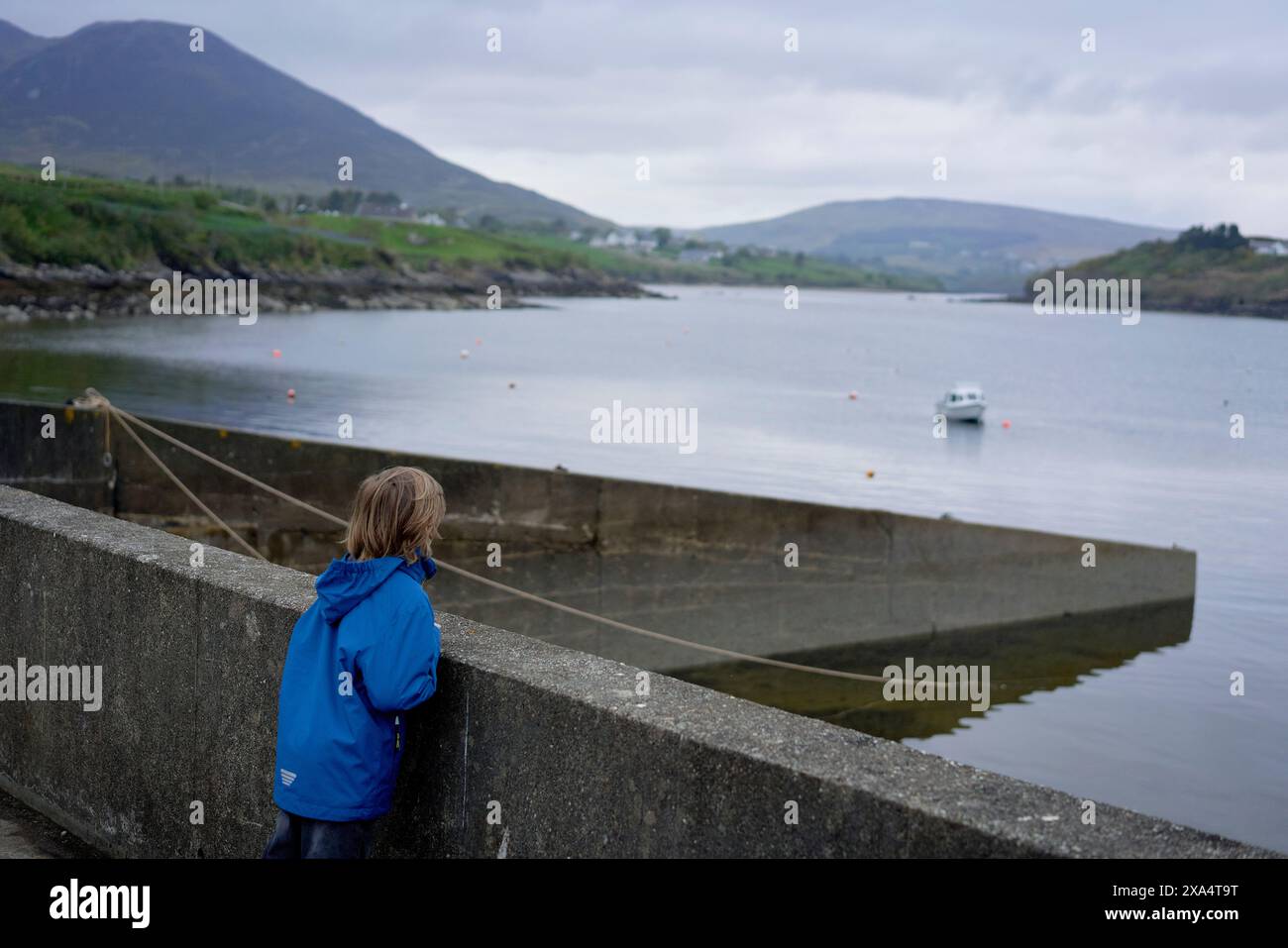 Kind in blauer Jacke mit Blick auf eine ruhige Bucht mit einem Boot und ferne Hügel unter einem bewölkten Himmel. Stockfoto