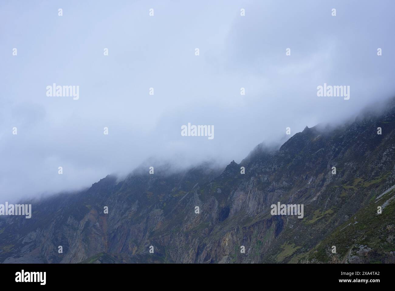 Neblige Berggipfel mit grauen Wolken, die die zerklüfteten Klippen umhüllen, und grüne Vegetation an den unteren Hängen. Stockfoto
