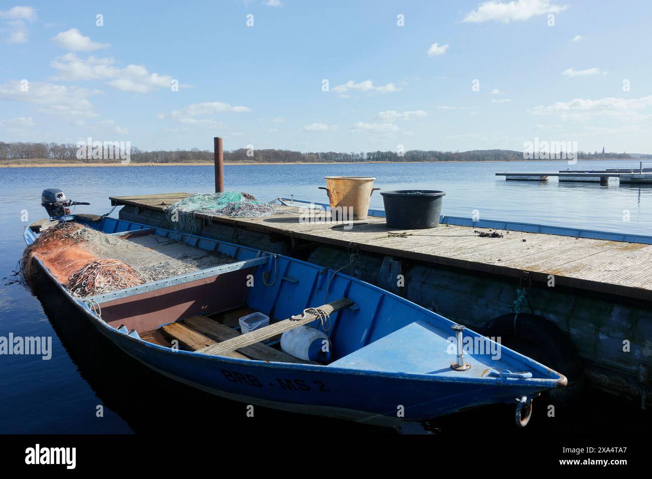 Ein blaues Fischerboot mit Netzen und Eimern, das an einem hölzernen Dock an einem ruhigen See unter teilweise bewölktem Himmel vertäut ist. Stockfoto