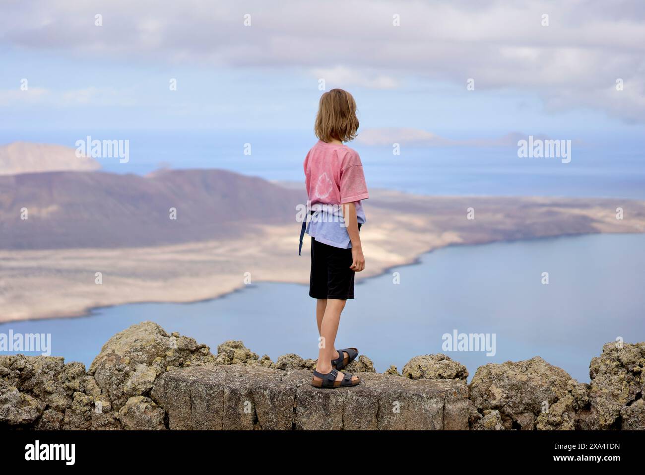 Eine Person steht auf einem felsigen Ausblick und blickt auf eine malerische Küstenlandschaft mit ruhigem blauem Wasser und fernen Inseln unter einem riesigen Himmel. Stockfoto
