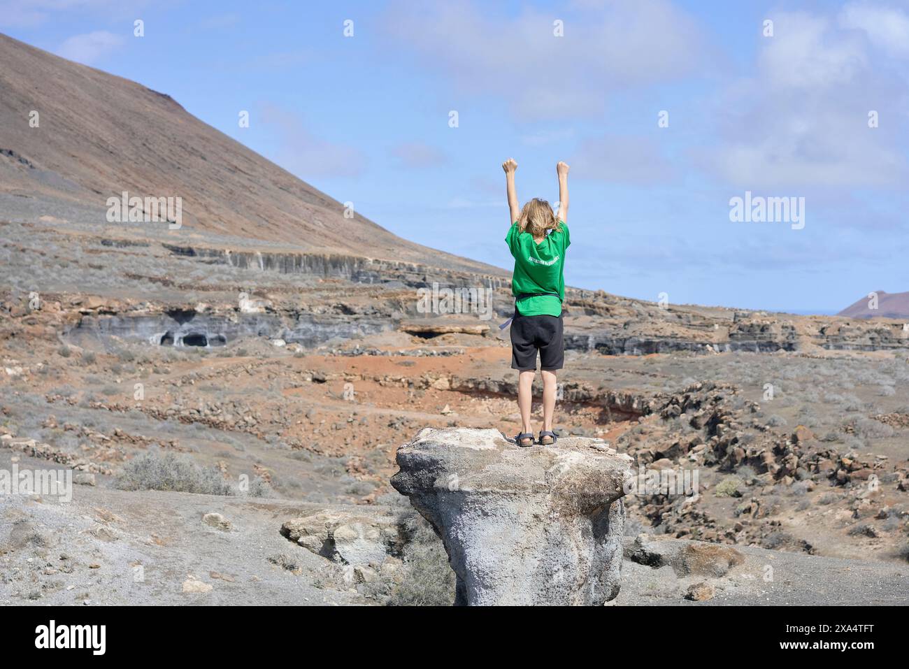 Ein Kind steht auf einem Felsen mit triumphalen Armen vor einer zerklüfteten Landschaft unter blauem Himmel. Stockfoto