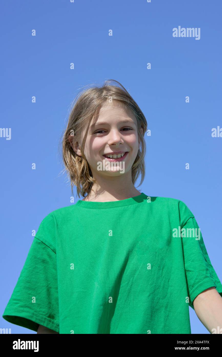 Lächelnder Junge in grünem Hemd, der vor einem klaren blauen Himmel steht. Stockfoto