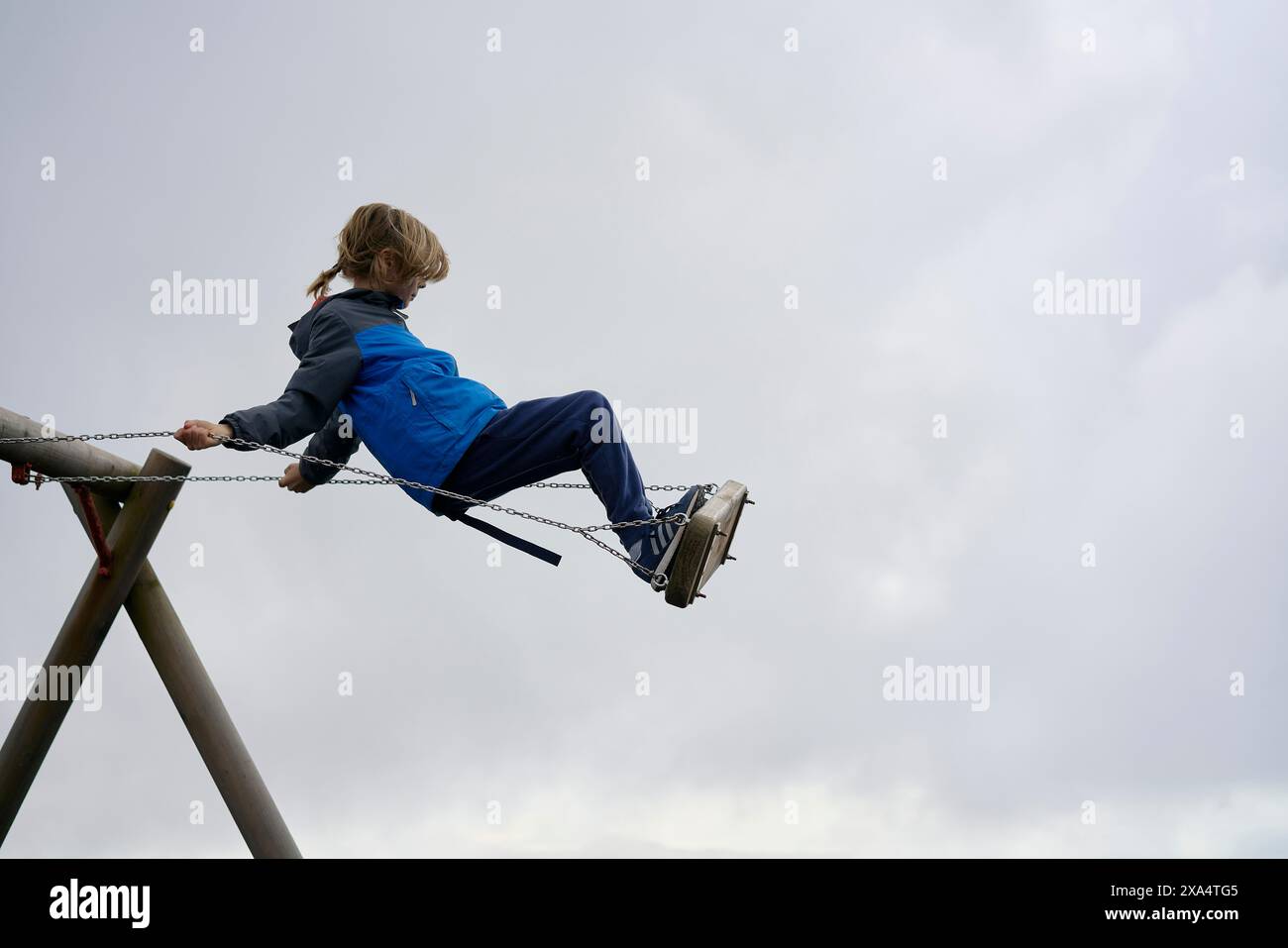 Ein Kind in einer blauen Jacke schwingt gerne hoch vor einem bewölkten Himmel. Stockfoto