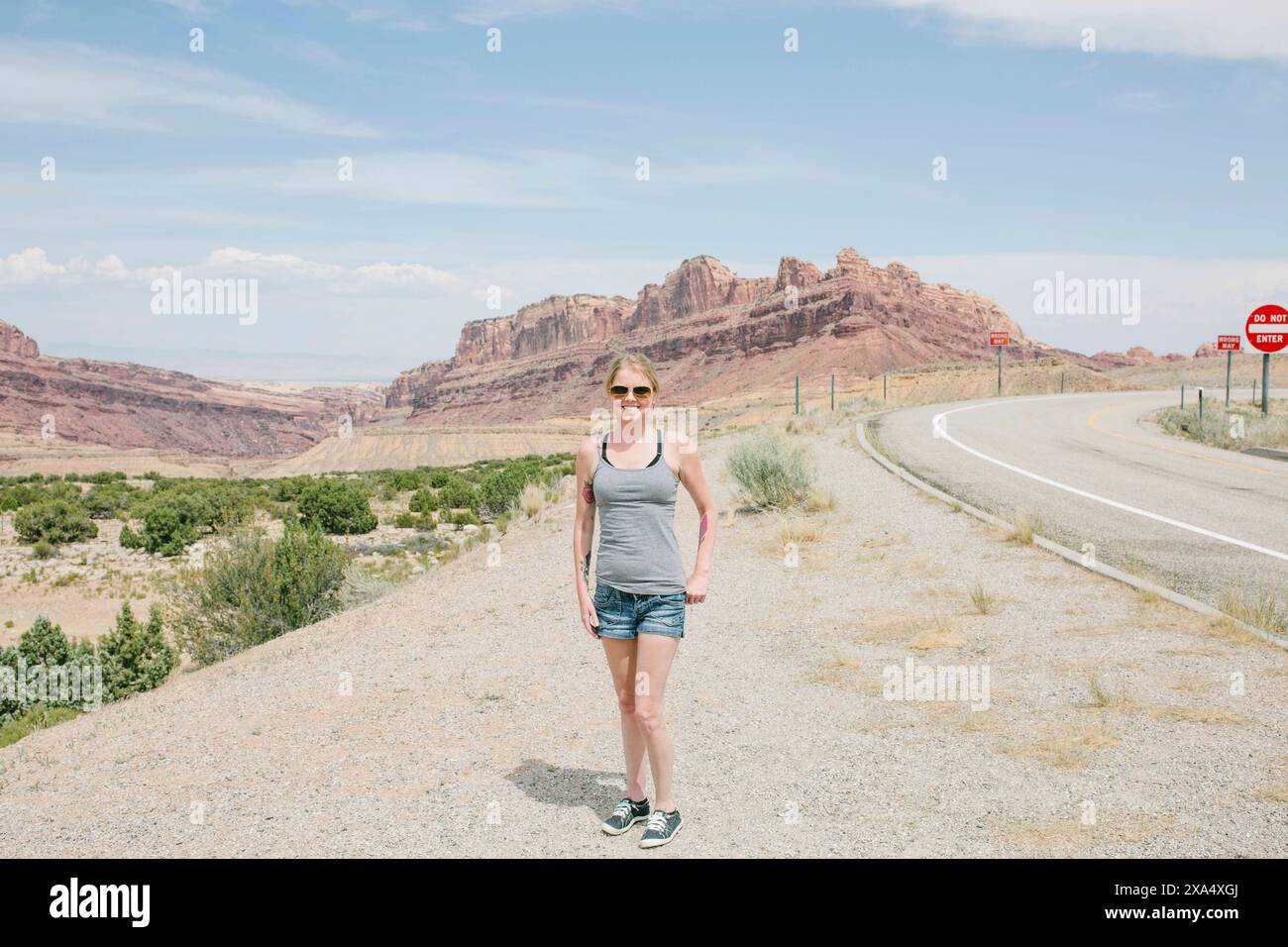 Lächelnde Frau in lässiger Kleidung posiert an einem sonnigen Tag am Straßenrand mit felsigem Wüstengelände im Hintergrund. Stockfoto