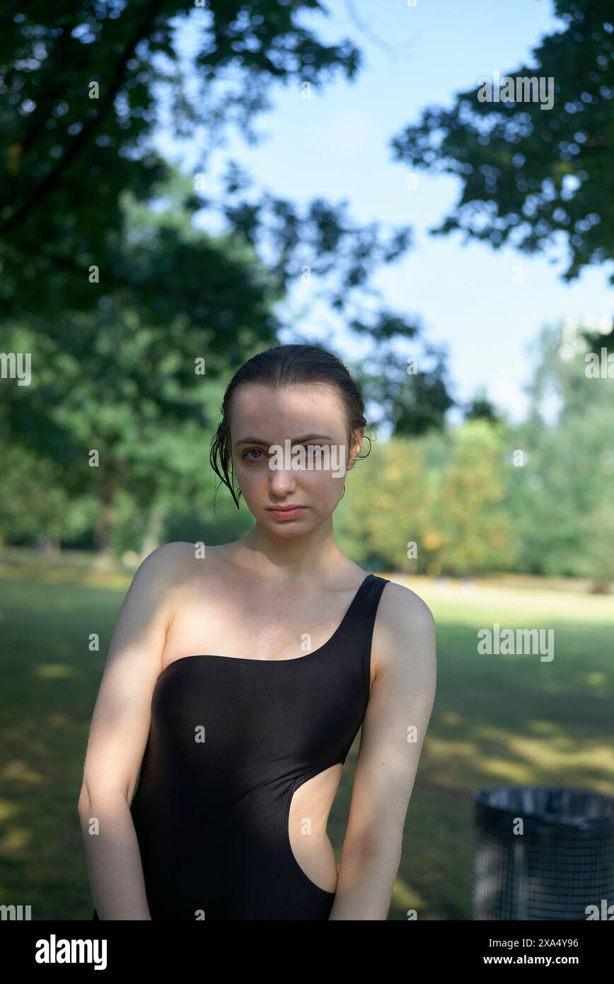 Frau in einem schwarzen ärmellosen Kleid, die an einem sonnigen Tag in einem Park mit Bäumen im Hintergrund steht. Stockfoto