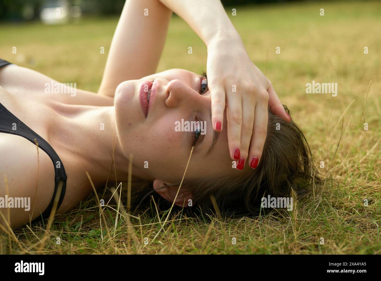 Eine junge Frau mit roten Fingernägeln, die auf Gras liegt und ihre Augen mit der Hand bedeckt. Stockfoto