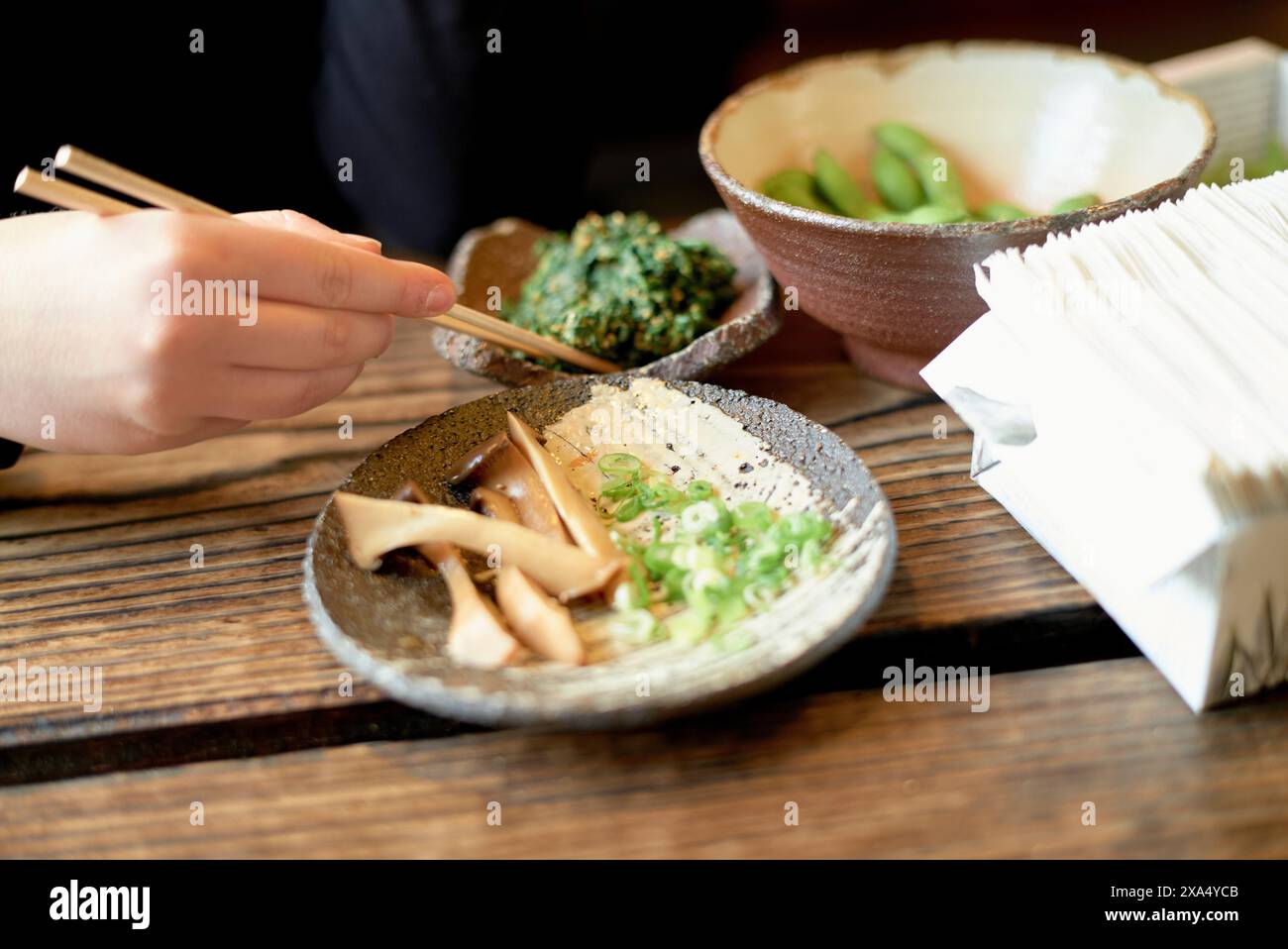 Mit der Hand die Stäbchen halten, um Lebensmittel aus einer Schüssel mit Beilagen auf einem Holztisch aufzuheben. Stockfoto