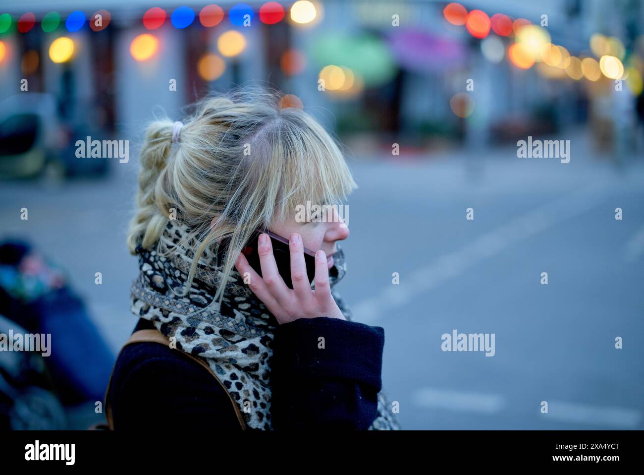 Eine Frau spricht mit ihrem Telefon, während sie in der Abenddämmerung auf einer Stadtstraße mit bunten Lichtern im Hintergrund läuft. Stockfoto