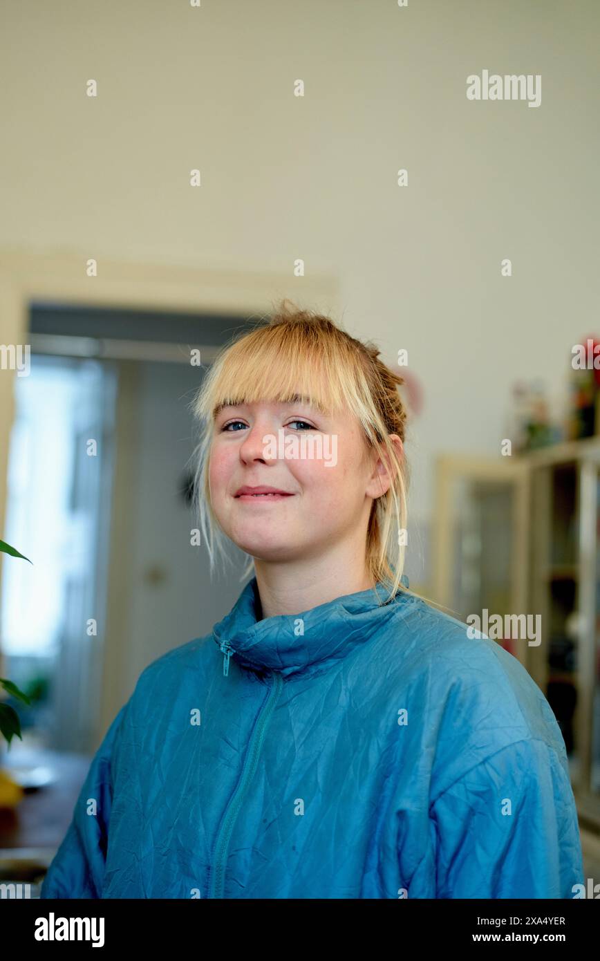 Lächelnde junge Frau mit blonden Haaren in einer blauen Jacke, die drinnen steht. Stockfoto