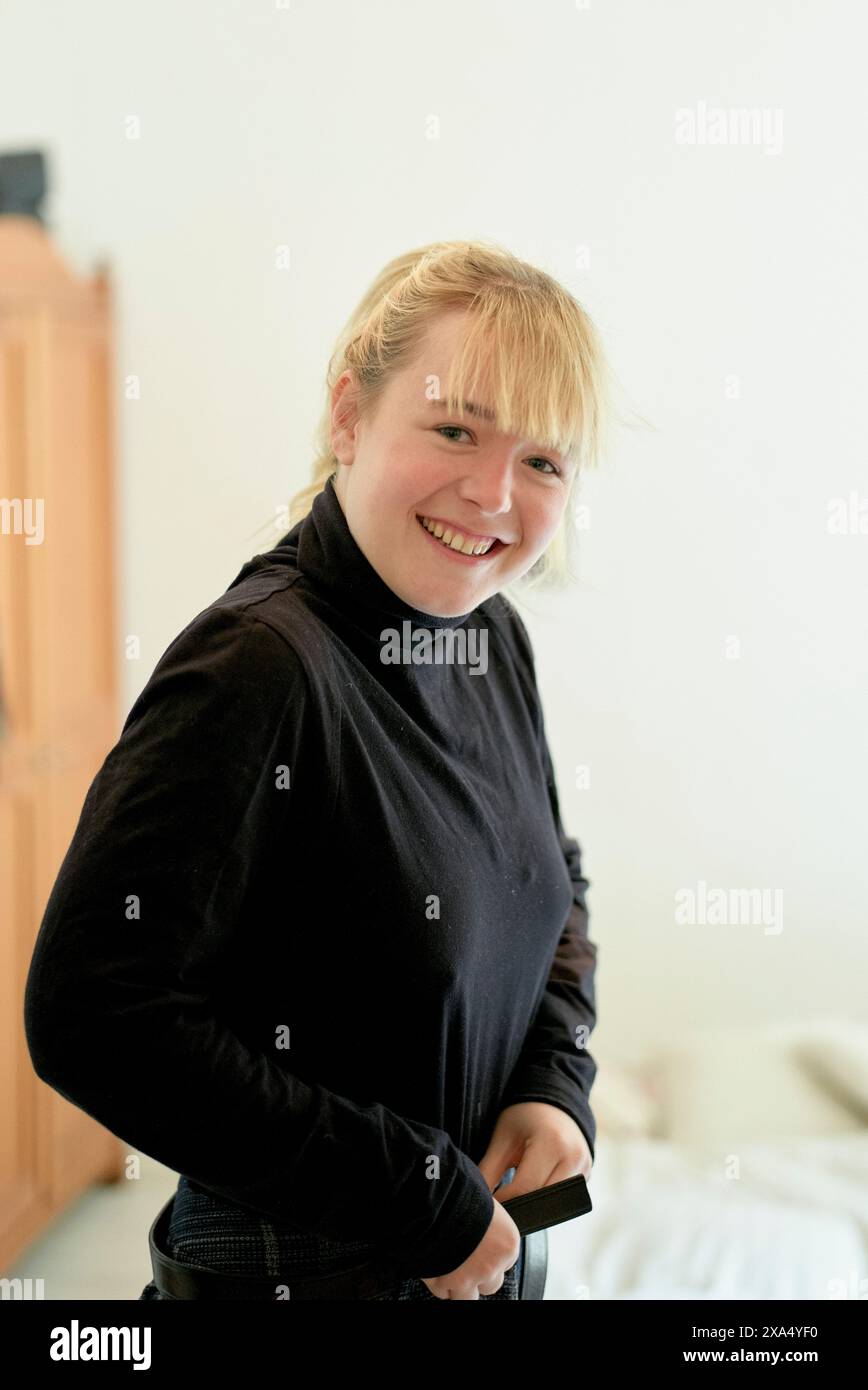 Lächelnde junge Frau in einem schwarzen Rollkragen, die drinnen mit neutralem Hintergrund steht. Stockfoto