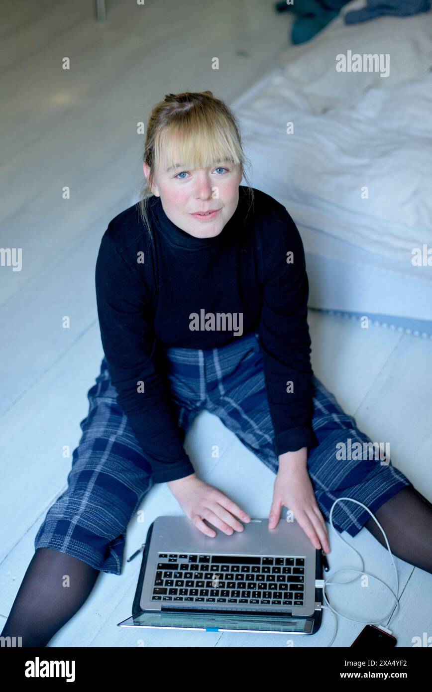 Eine Frau sitzt auf dem Boden mit einem Laptop auf dem Schoß und schaut in die Kamera. Stockfoto