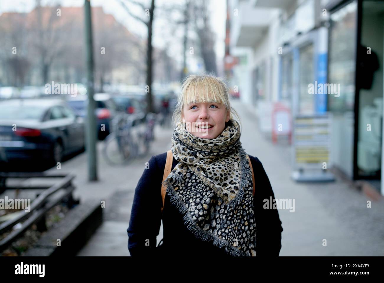 Lächelnde Frau mit blonden Haaren, die einen gemusterten Schal trägt, steht auf einer Stadtstraße mit Bäumen und geparkten Autos im Hintergrund. Stockfoto