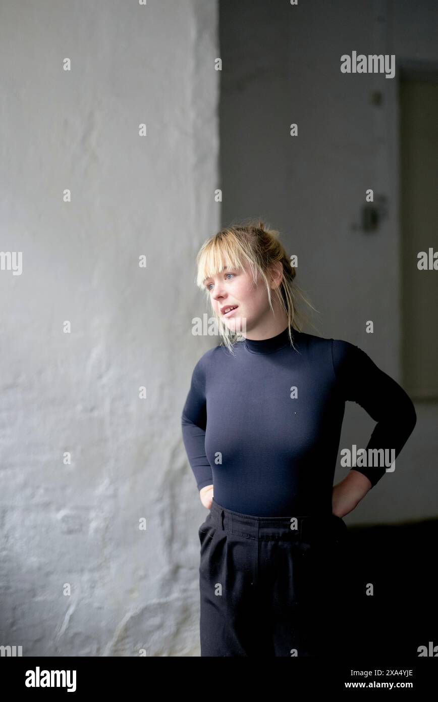 Eine selbstbewusste Frau mit blonden Haaren im Pferdeschwanz, marineblauem Oberteil und schwarzer Hose, die mit einer reflektierenden Pose vor einer weißen Wand steht. Stockfoto
