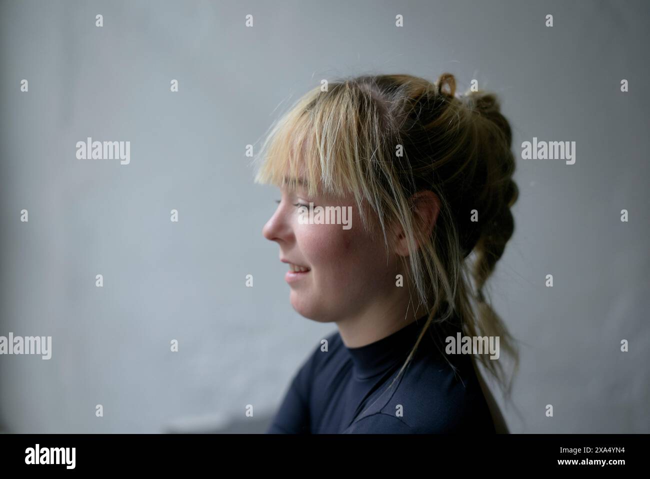 Seitenprofil einer lächelnden jungen Frau mit blondem Haar vor grauem Hintergrund. Stockfoto