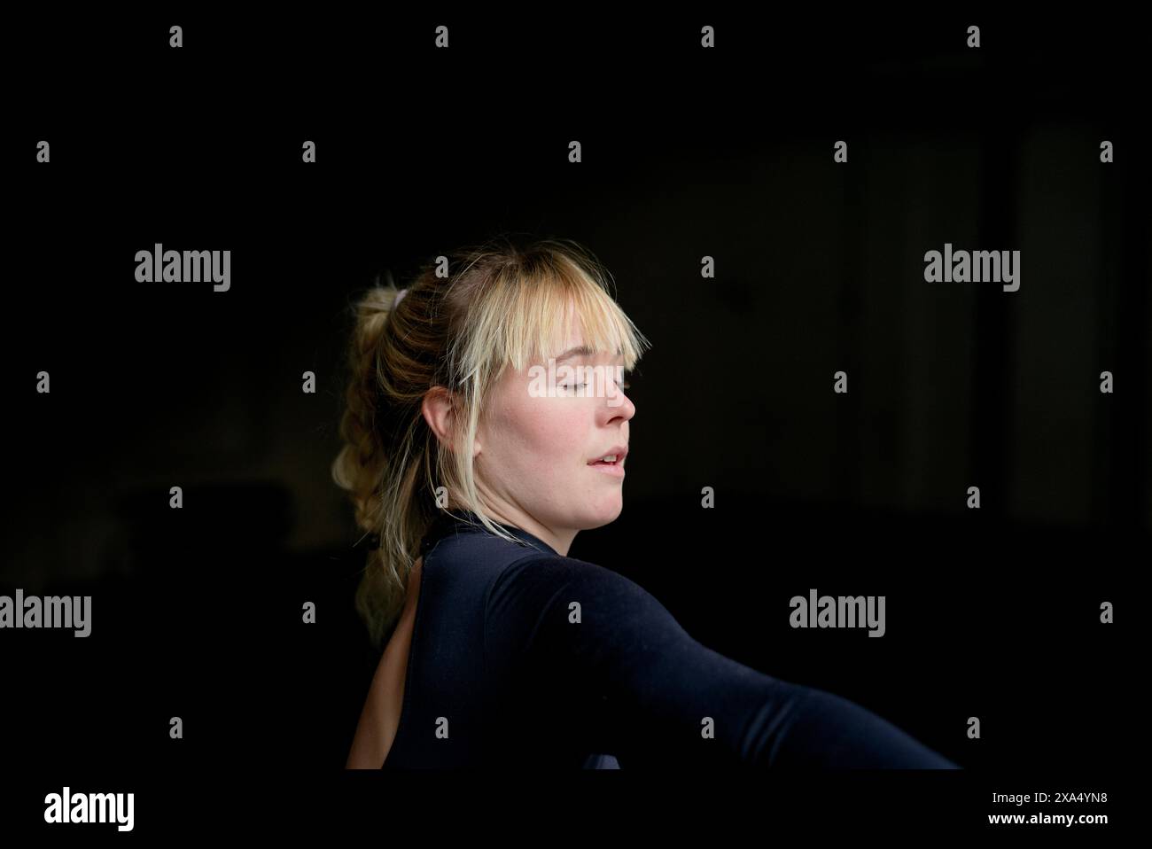 Eine junge Frau mit den Haaren in einem lässigen Brötchen, geschlossenen Augen und hinter dem Rücken gestreckten Armen in einer friedlichen Haltung vor dunklem Hintergrund. Stockfoto