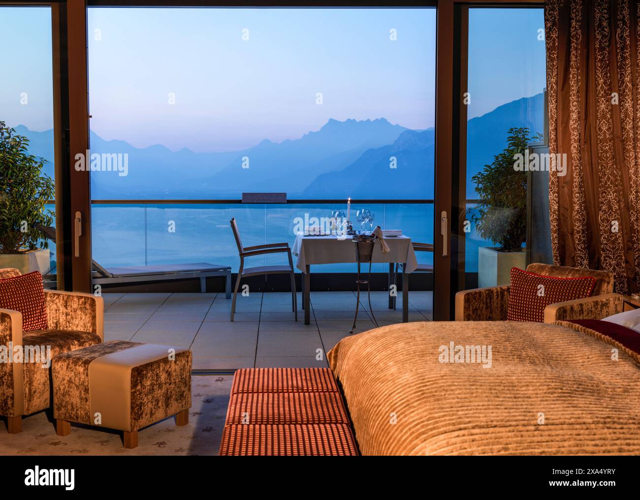 Elegantes Hotelzimmer mit Blick auf eine Bergkette in der Dämmerung, einem Plüschbett, einem Sitzbereich und einem Essbereich auf dem Balkon. Stockfoto