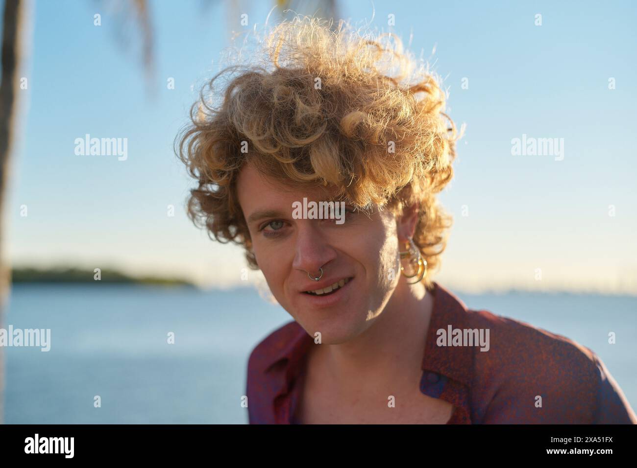 Lockiger junger Erwachsener mit Nasenring lächelt sanft vor dem Hintergrund einer ruhigen Skyline und Wasser während der goldenen Stunde. Stockfoto