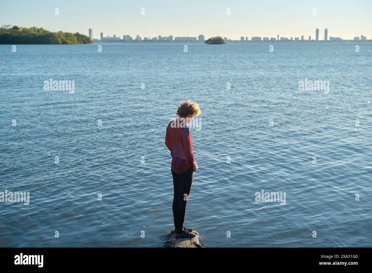 Frau, die auf einem Felsen am Rande eines ruhigen Sees steht und bei Tageslicht zur Skyline blickt. Stockfoto