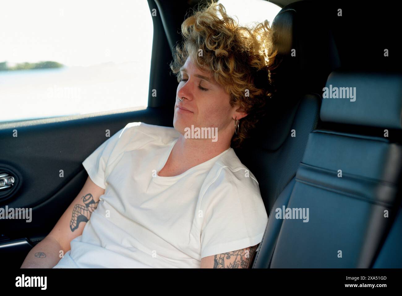 Junger erwachsener Mann mit lockigen Haaren und Tätowierungen, der auf dem Rücksitz eines Autos schläft. Stockfoto