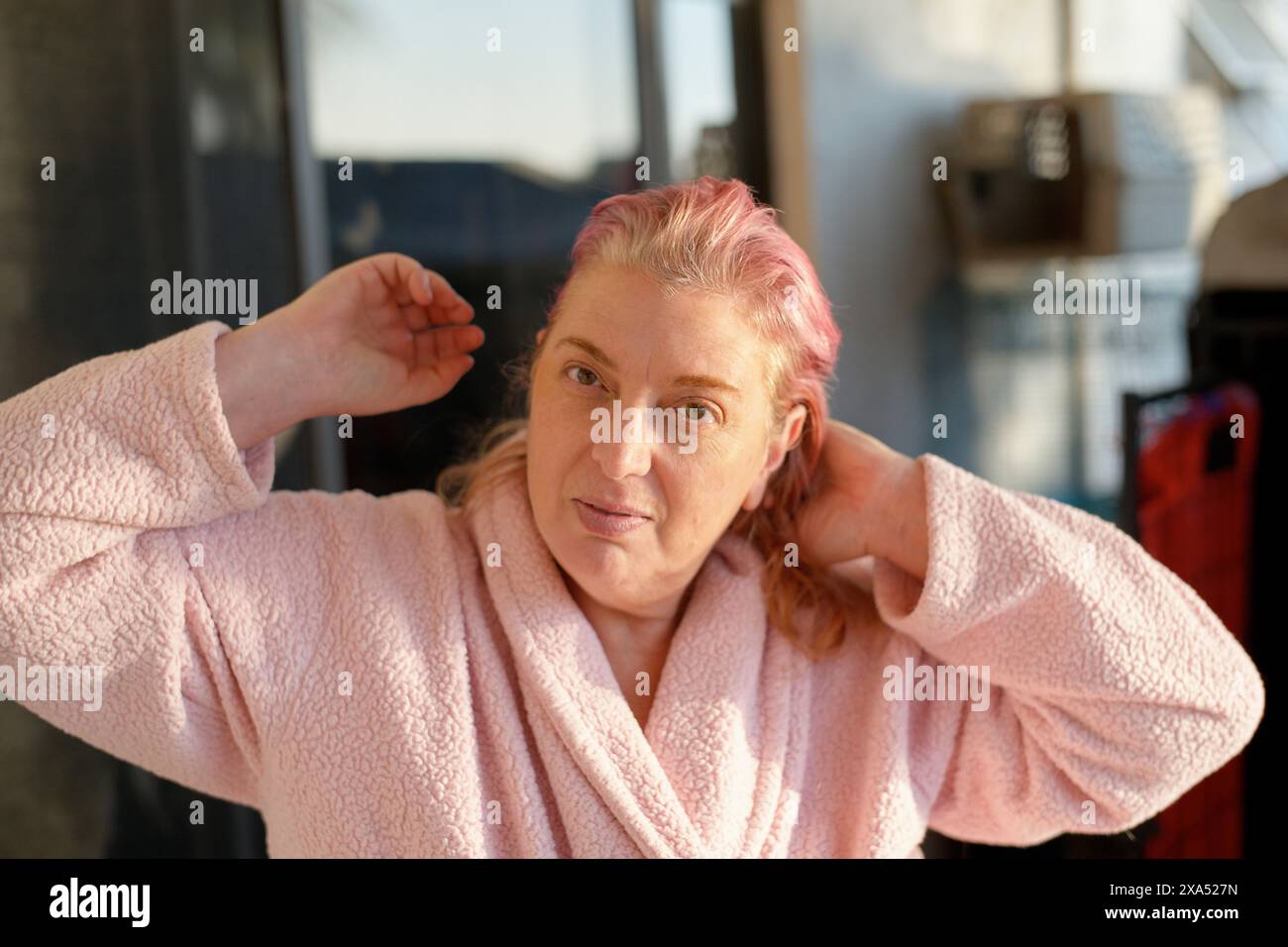 Eine Frau mittleren Alters mit rosafarbenen Haaren bindet ihre Haare zurück, während sie in einem sonnendurchfluteten Zimmer einen rosafarbenen Bademantel trägt. Stockfoto