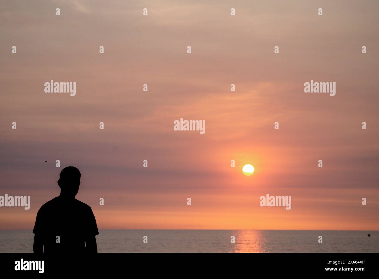 Ein Mann steht am Sandstrand und blickt auf den Horizont, wo die orangen Sonnenuntergänge auf das Meer treffen. Es ist ein Moment der Reflexion. Stockfoto