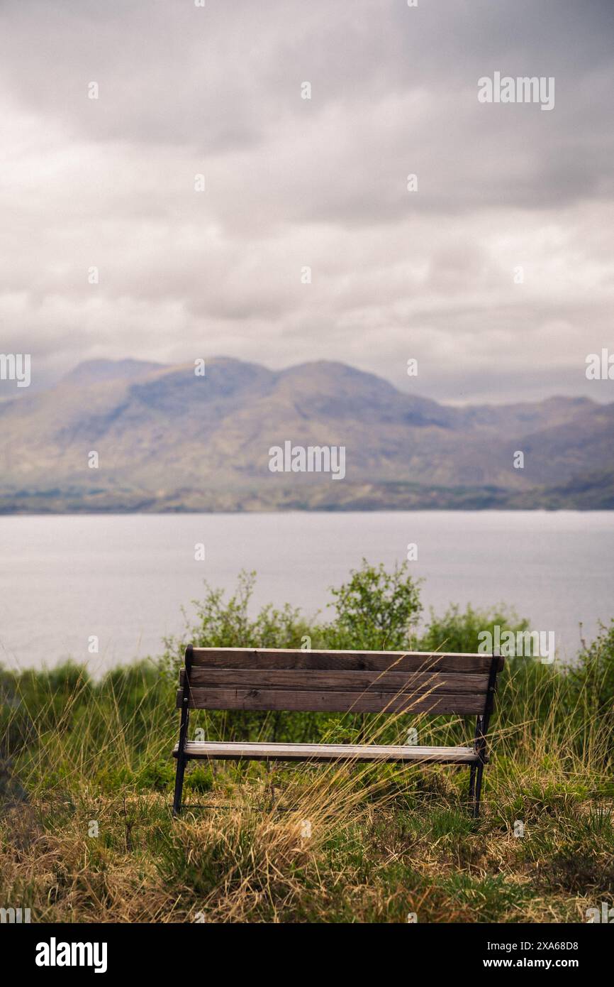 Eine Holzbank mit Blick auf einen bewölkten See und Hügel. Stockfoto