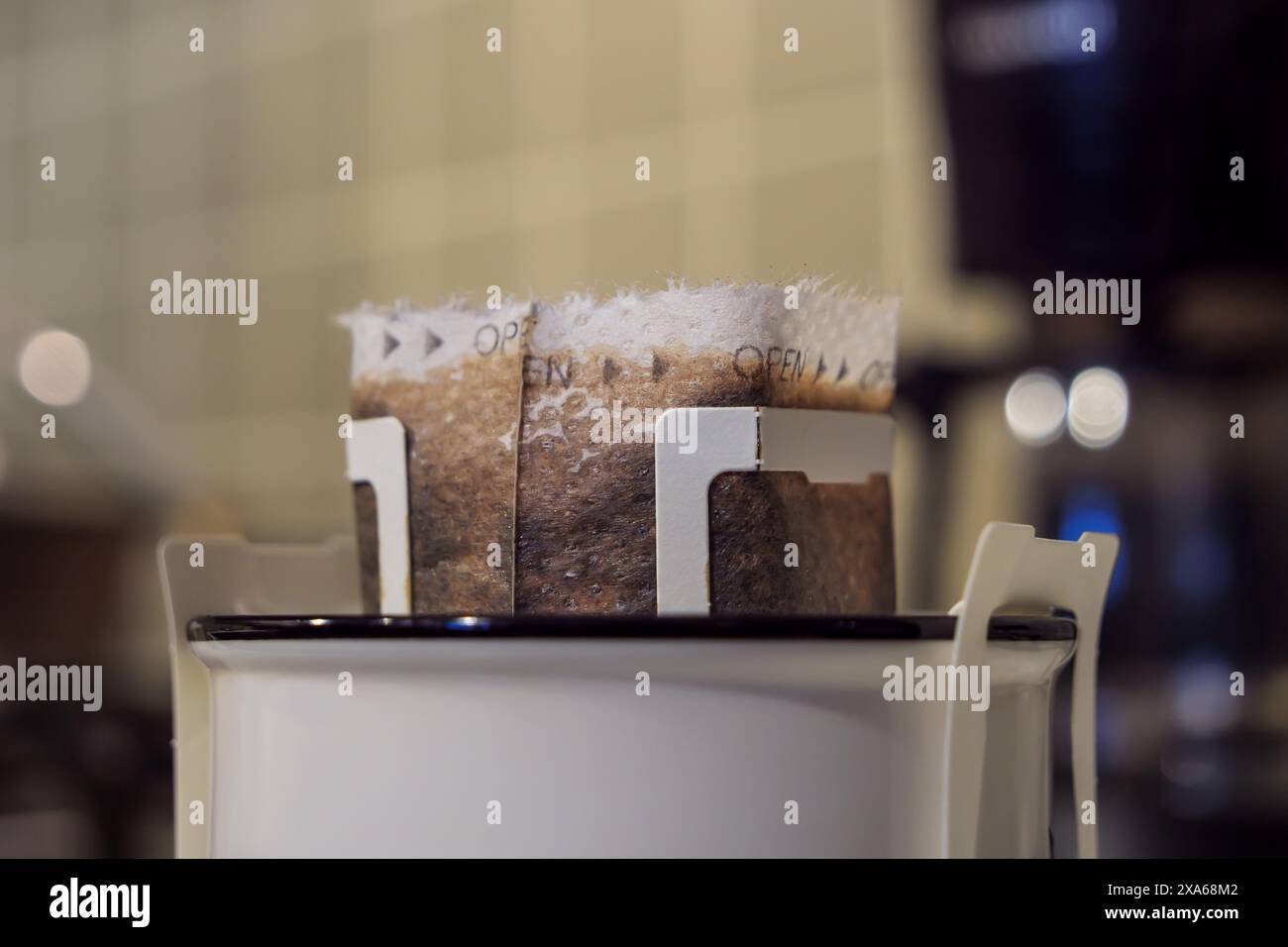 Ein Kaffeebeutel mit Tropffilter wird offen in einer Keramikschale vor einem verschwommenen Küchenhintergrund platziert, bereit zum Brühen, extrem aus nächster Nähe. Stockfoto
