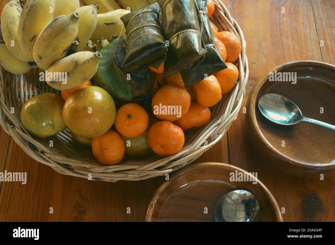 Ein Korbkorb gefüllt mit tropischen Sommerfrüchten wie Passionsfrüchten, Bananen und Orangen auf einem Holztisch neben leeren Holzschalen mit Löffel Stockfoto