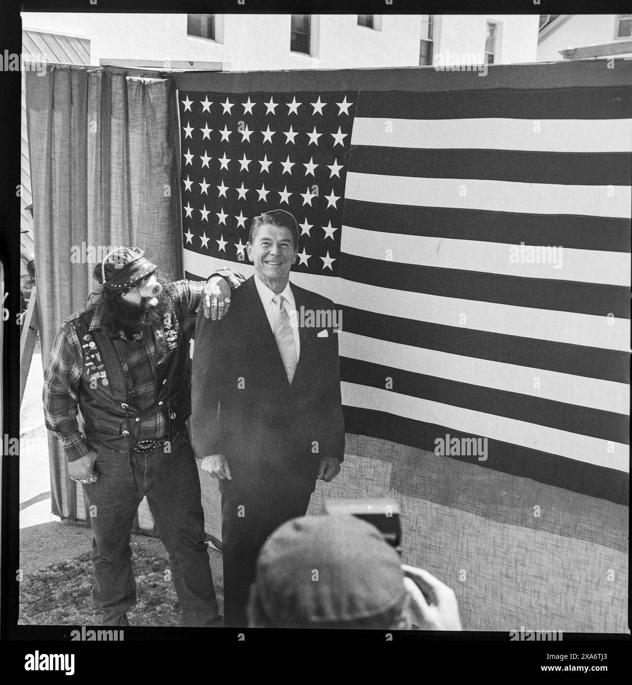 Biker mit aufgesetztem Schweinerüssel als Nase posiert mit Pappfigur von Präsident Ronald Reagan vor amerikanischer Flagge während der Daytona Bike Week in Daytona Beach, Florida USA am 15.3.1985 *** Biker mit Schweineschnauze für eine Nase posiert mit einem Pappausschnitt von Präsident Ronald Reagan vor der amerikanischen Flagge während der Daytona Bike Week am 15. März 1985 in Daytona Beach, Florida USA Stockfoto