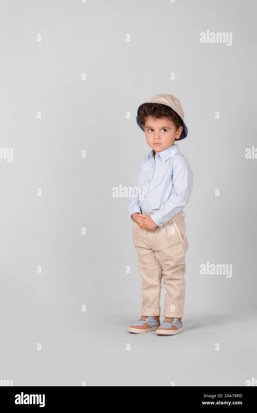 Ein kleiner Junge in hellbraunen Hosen und einem braunen Hut steht auf einem weißen Hintergrund Stockfoto