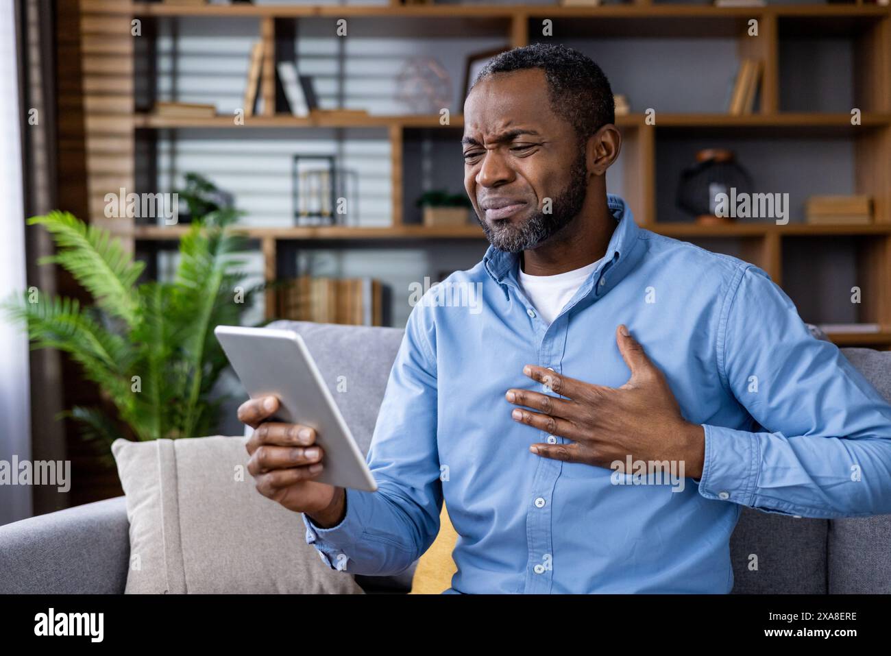 Ein kranker Patient hält seine Brust, während er sich mit einem Arzt über einen Videoanruf aus seinem Wohnzimmer berät. Gesundheitswesen und Telemedizin-Konzept. Stockfoto