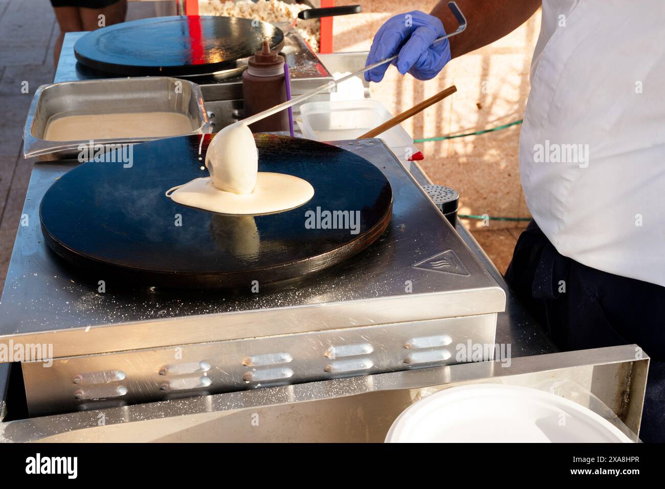 Ein Mann macht Pfannkuchen auf einer Grillplatte. Eine Frau hält ein Baby in einem Kinderwagen Stockfoto