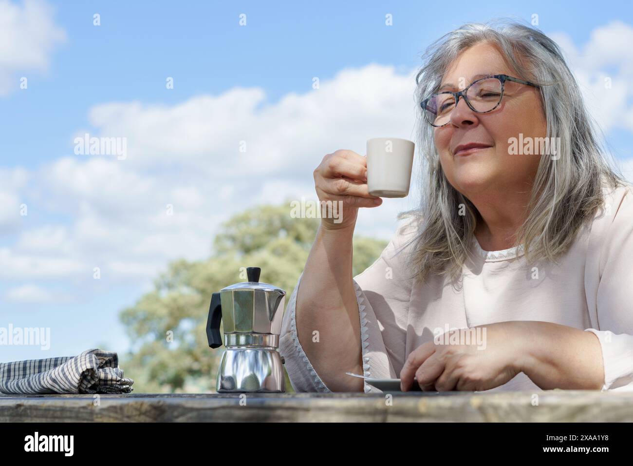 Nahaufnahme einer älteren Frau mit weißem Haar, die über einer Tasse Kaffee an einem Tisch auf einem Feld lächelt, mit einem bewölkten blauen Himmel im Hintergrund Stockfoto