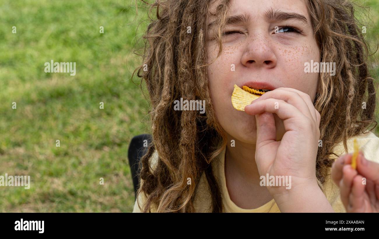 Genießen Sie den Geschmack der Kindheit in dieser Nahaufnahme: Ein blonder Junge mit Dreadlocks genießt einen Snack im Park. Freude und Entspannung verschmelzen, während er sich verwöhnt Stockfoto