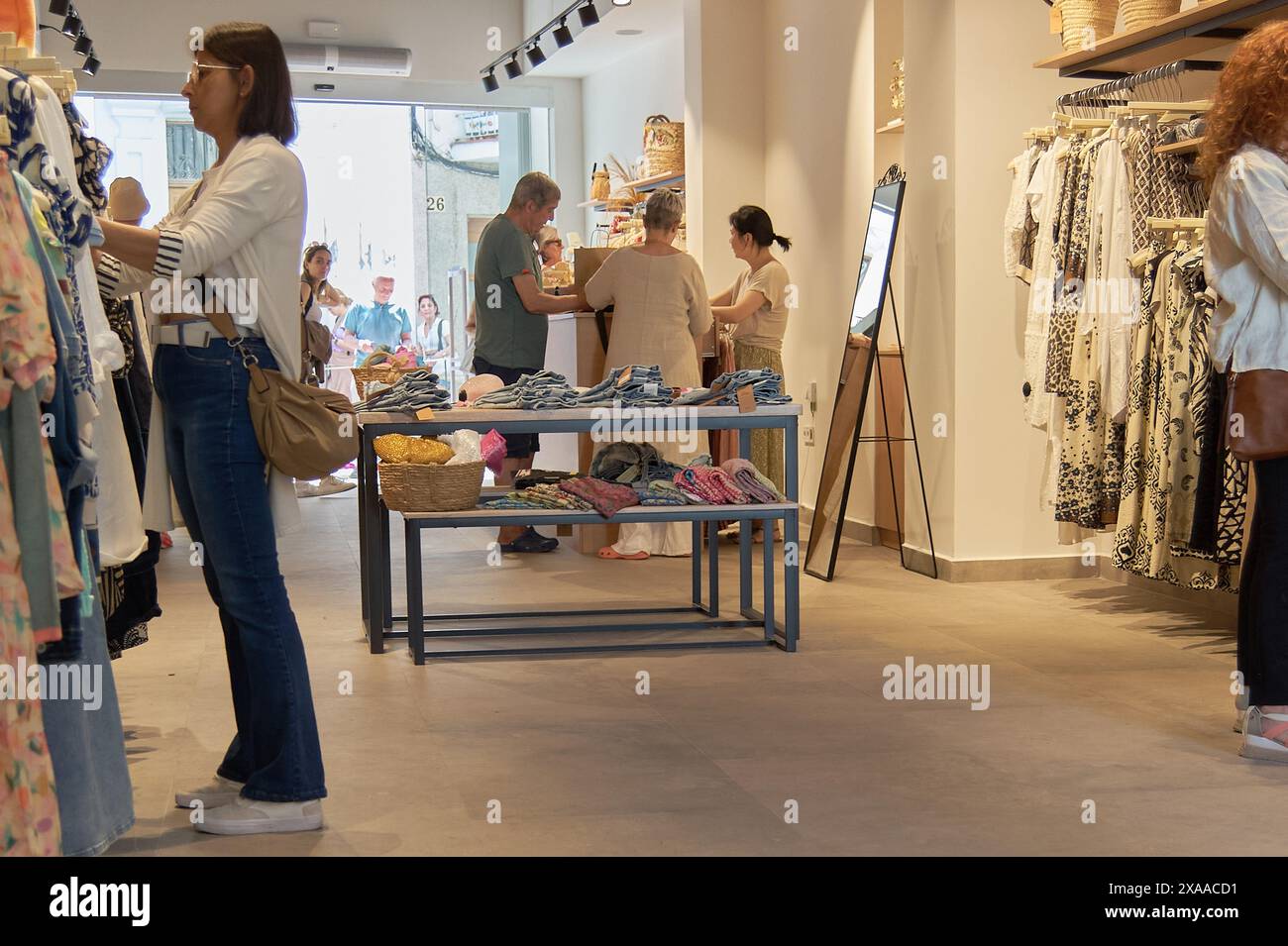 Sitges, Barcelona, Spanien – 05. Juni 2024: Kunden wählen Kleidung in einem modernen Geschäft aus, das mit verschiedenen Kleidungsstücken auf Regalen und Tischen gefüllt ist. Einkaufsmöglichkeiten am Vormittag Stockfoto