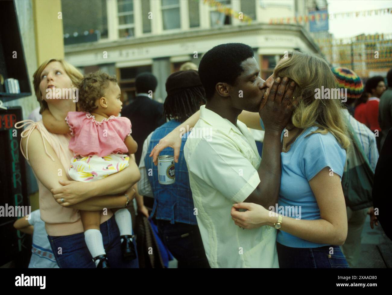 Der junge britische Schwarze britische Mann der 1970er Jahre küsst seinen weißen britischen Partner, hinter ihnen hält ein Freund sein Baby mit gemischter Rasse. Notting Hill Carnival, London, England, 27. August 1979. HOMER SYKES Stockfoto