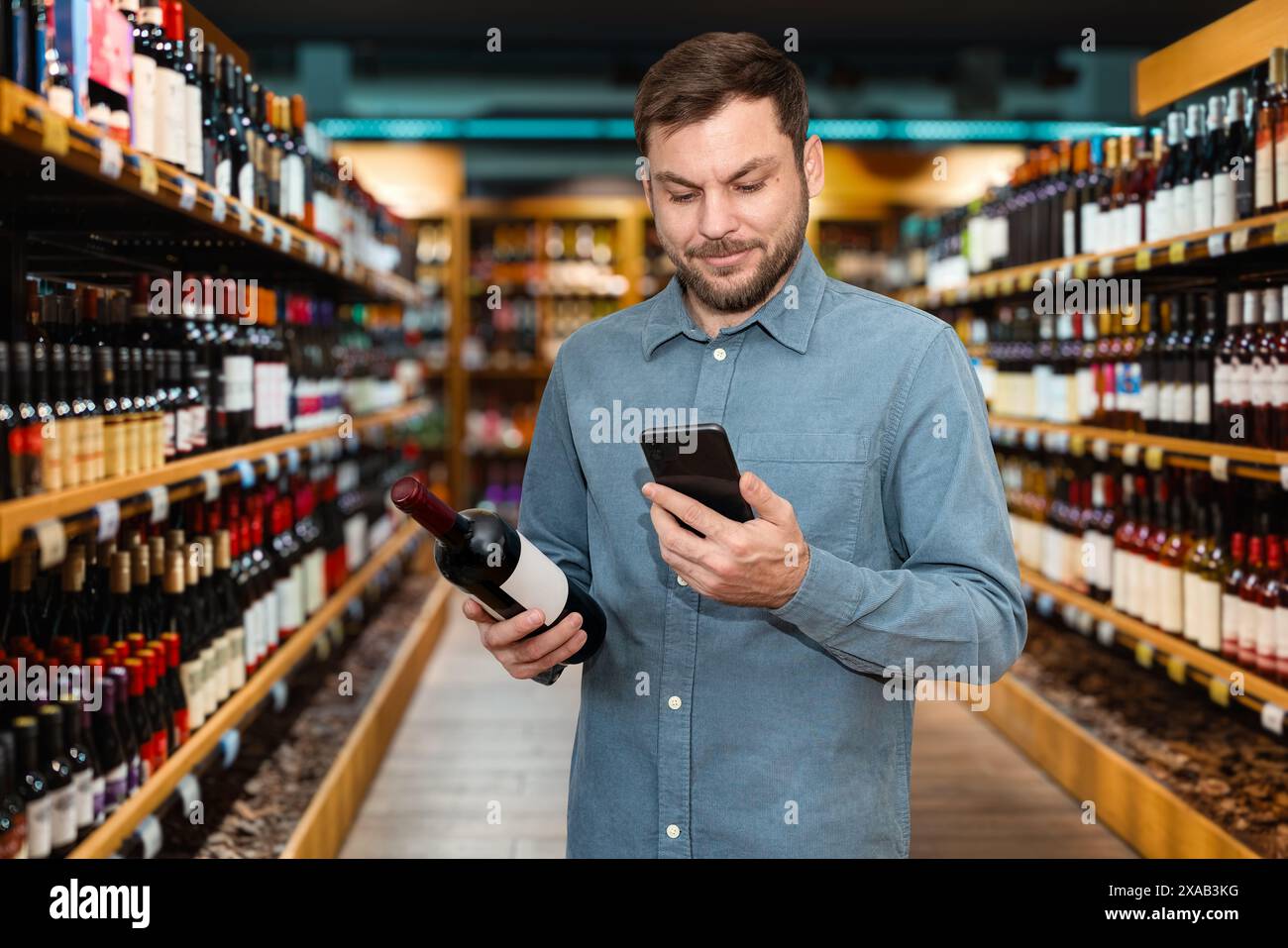Schöner Mann, der Wein im Spirituosenladen auswählt. Männlicher Kunde hält Rotweinflasche und Handy in der Hand, während er in einem Weinladen einkaufen kann. Stockfoto