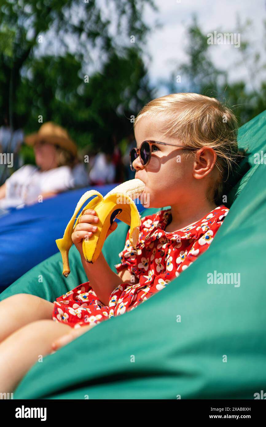 Ein kleines Mädchen isst eine Banane. Porträt eines modischen, stilvollen, schönen Mädchens in Sonnenbrille und rotem Kleid, während er in einem Sitzsack-Stuhl sitzt Stockfoto