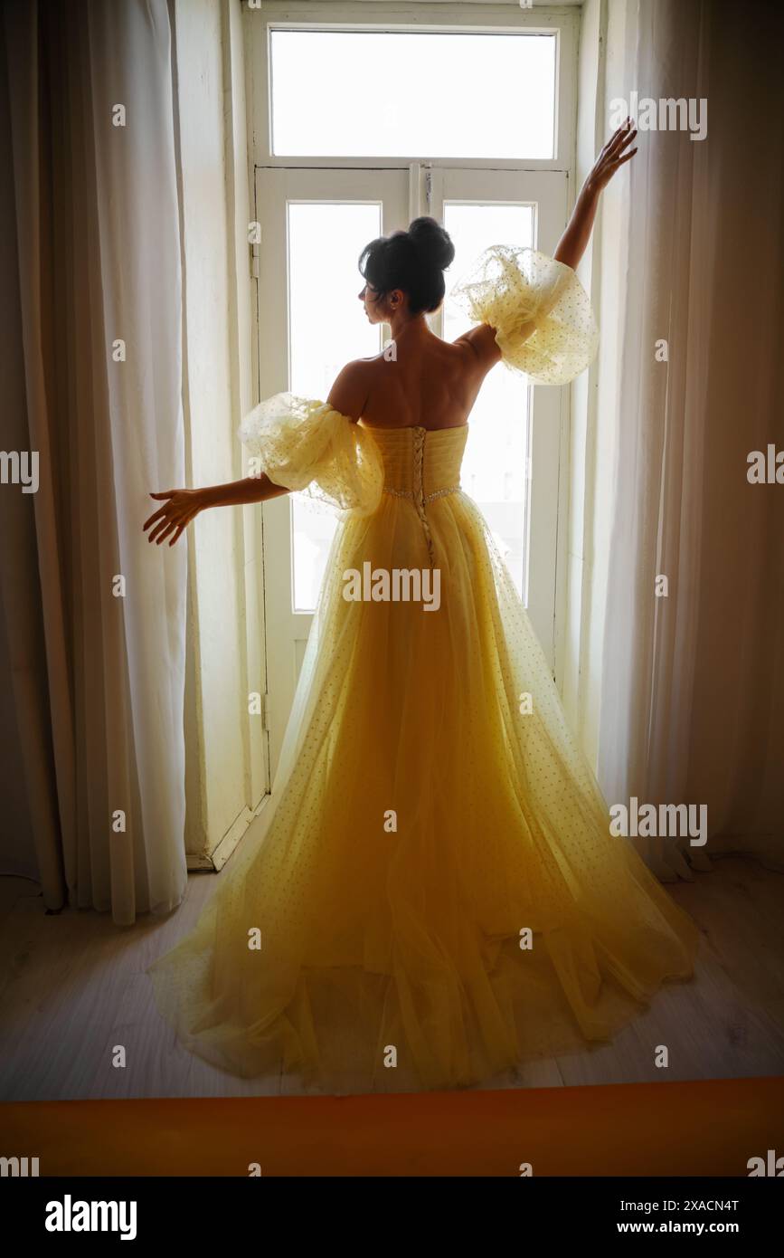 Die Silhouette einer Frau in einem goldenen, luxuriösen Kleid vor dem Hintergrund eines Fensters hält einen Vorhang mit den Händen. Elegante Dame in gelber Seide Stockfoto