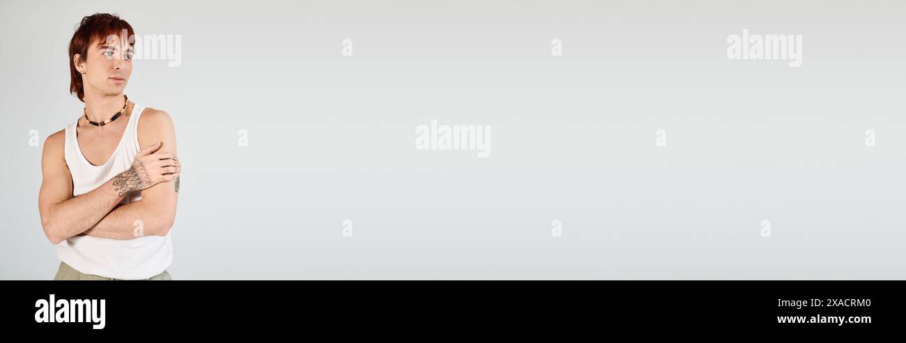 Ein stilvoller junger Mann posiert selbstbewusst in einem weißen Tanktop und steht mit gekreuzten Armen in einem Studio mit grauem Hintergrund. Stockfoto