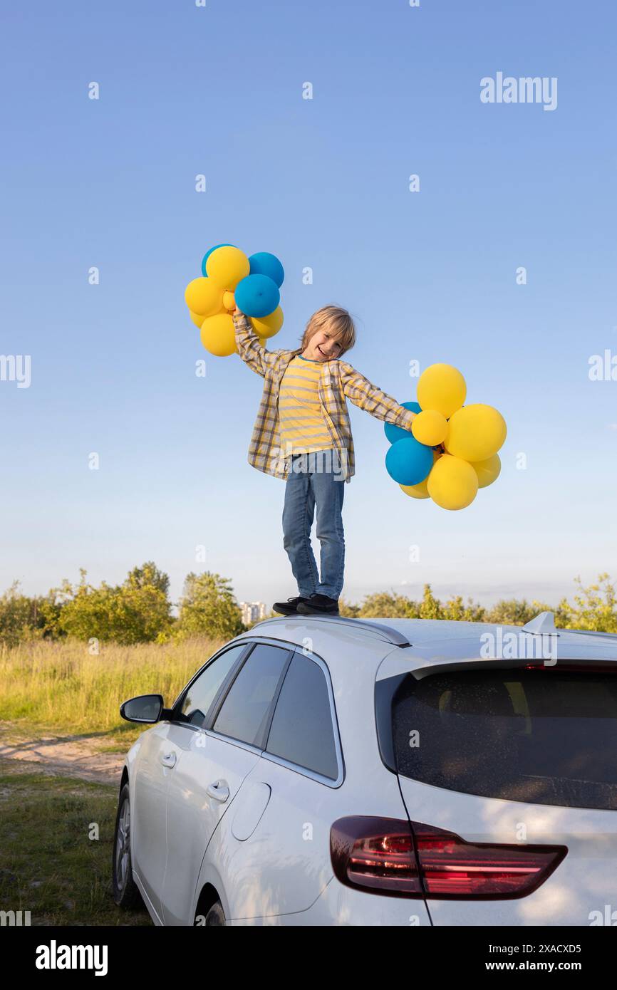 Glücklicher Junge, der auf dem Dach des Autos steht und viele gelbe und blaue Ballons am Himmel hält. Fühlt sich toll an, Urlaub, Geburtstag. Unabhängigkeitstag der Ukraine Stockfoto