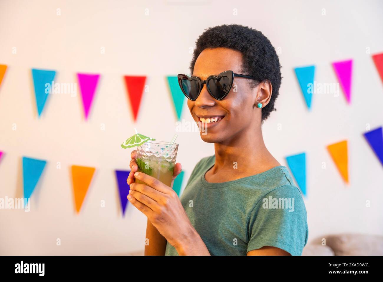 Porträt eines glücklichen lateinischen Mannes mit Sonnenbrille, der Cocktail neben Girlanden trinkt, die ein Haus während der Feier schmücken Stockfoto