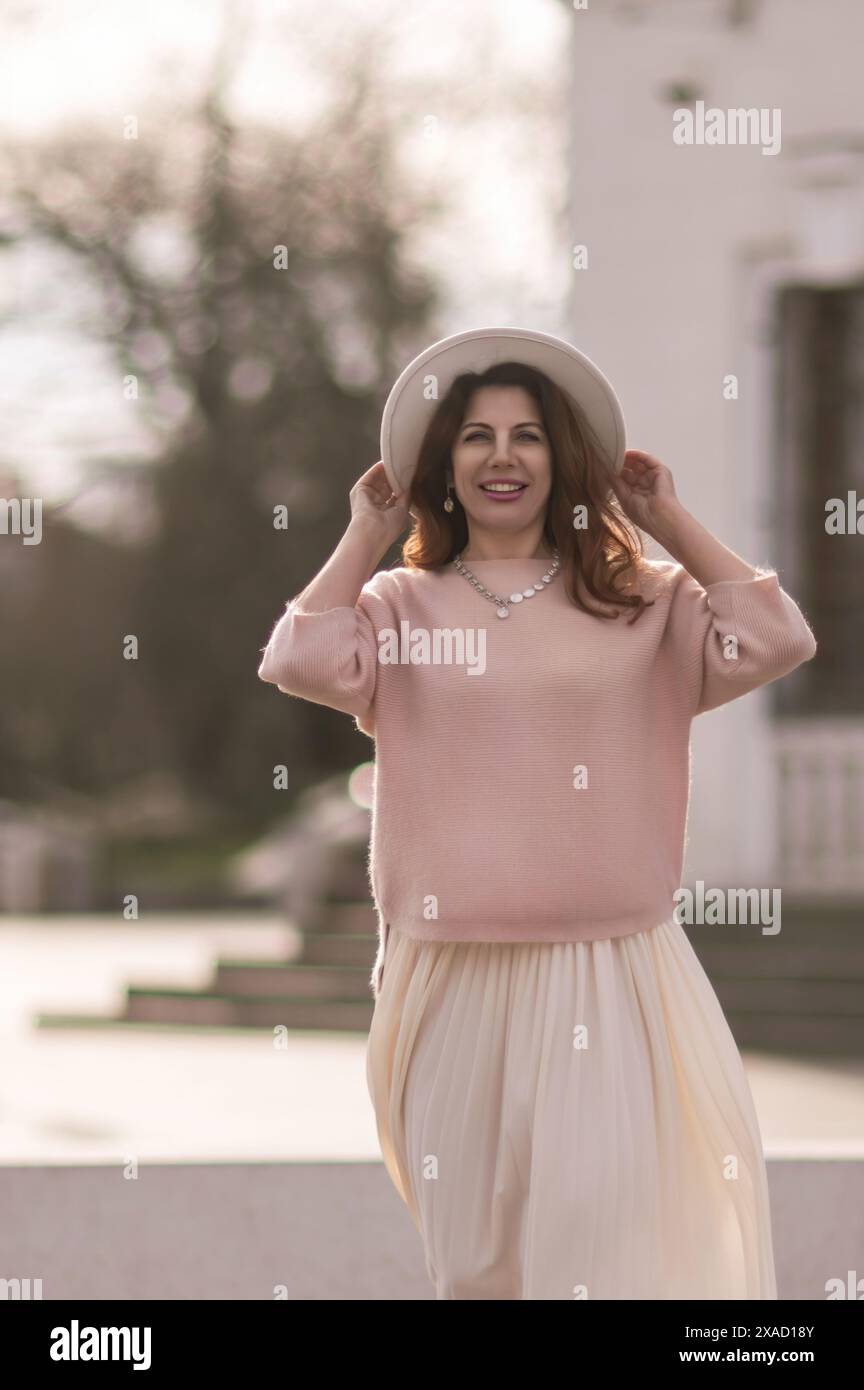 Eine Frau mit rosa Pullover und weißem Rock lächelt und hält ihren Hut. Das Bild hat eine warme und fröhliche Stimmung, mit dem Lächeln der Frau und dem Stockfoto