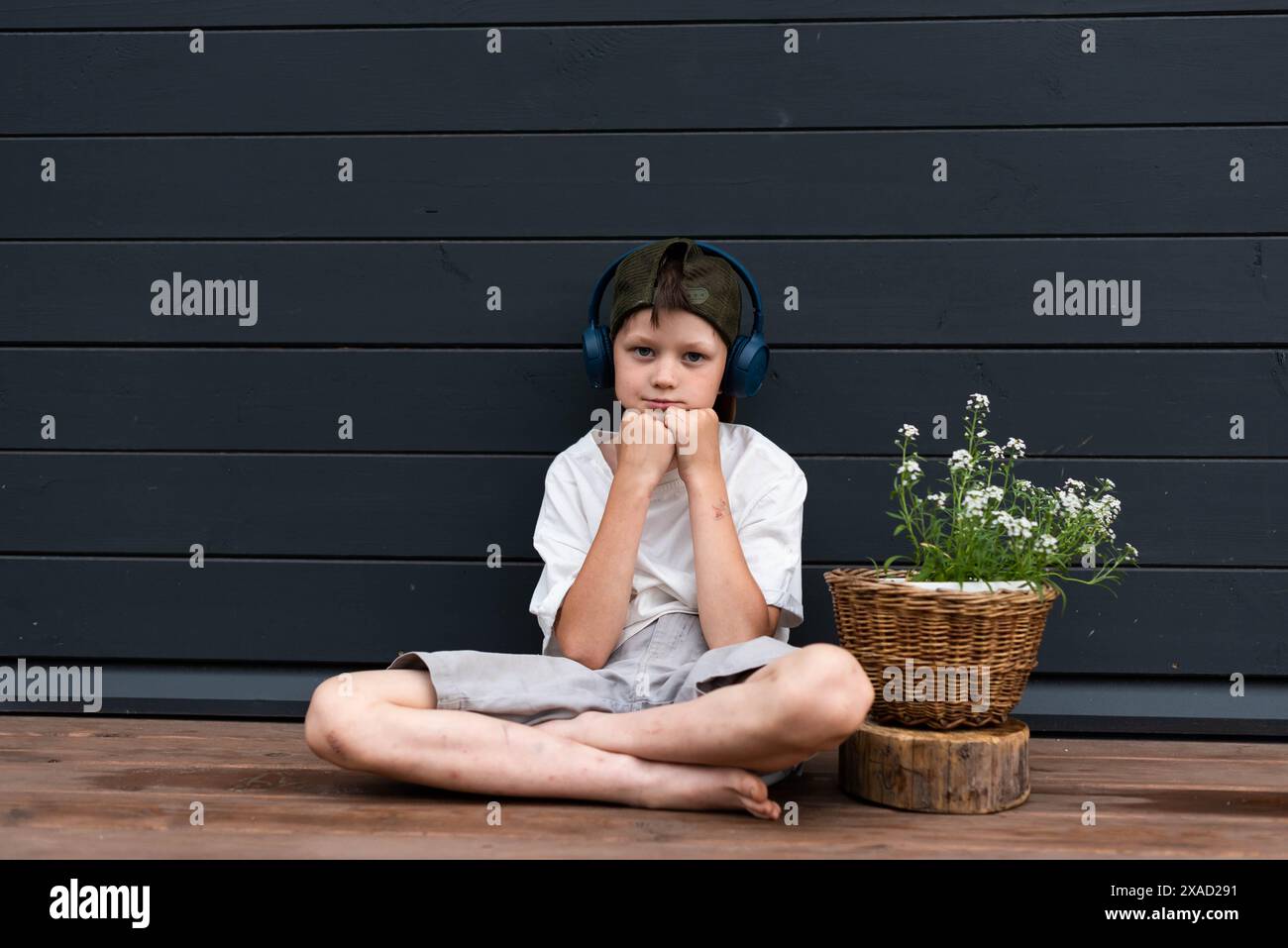 Geballter Jugendlicher in Kopfhörern, der auf einer Holzterrasse in der Nähe einer Blume sitzt Stockfoto